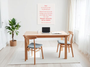 Helles, minimalistisches Arbeitszimmer mit Holztisch und zwei Stühlen. Auf dem Tisch stehen eine Tasse, ein aufgeklapptes Notizbuch und ein Laptop. Links neben dem Tisch steht eine grüne Zimmerpflanze im Korb. An der Wand hängt ein Bild mit dem Zitat: „Mut ist nicht immer brüllend laut. Manchmal ist es die ruhige, leise Stimme am Ende des Tages, die sagt: Morgen versuche ich es wieder.“