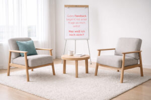 Heller Coachingraum mit zwei grauen Sesseln auf einem hellen Teppich und einem runden Holztisch mit Tasse und Notizbuch. Im Hintergrund steht ein Flipchart mit der Aufschrift: „Gutes Feedback beginnt mit einer Frage an mich selbst: Was weiß ich noch nicht?“